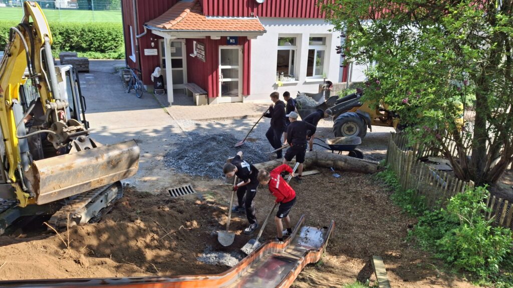 Ein Bagger und sechs Schüler graben den Bereich um eine Rutsche um.