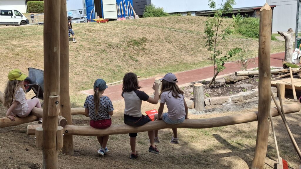 Vier Mädchen sitzen auf Holzbalken vor der Mitmachbaustelle.
