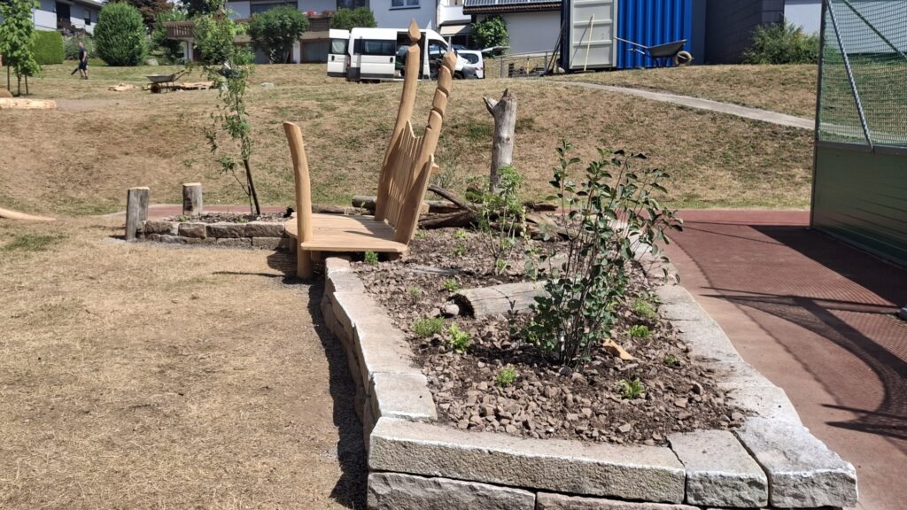 Sitzgelegenheit aus Holz vor entsiegelter und bepflanzter Fläche vor dem Sportplatz