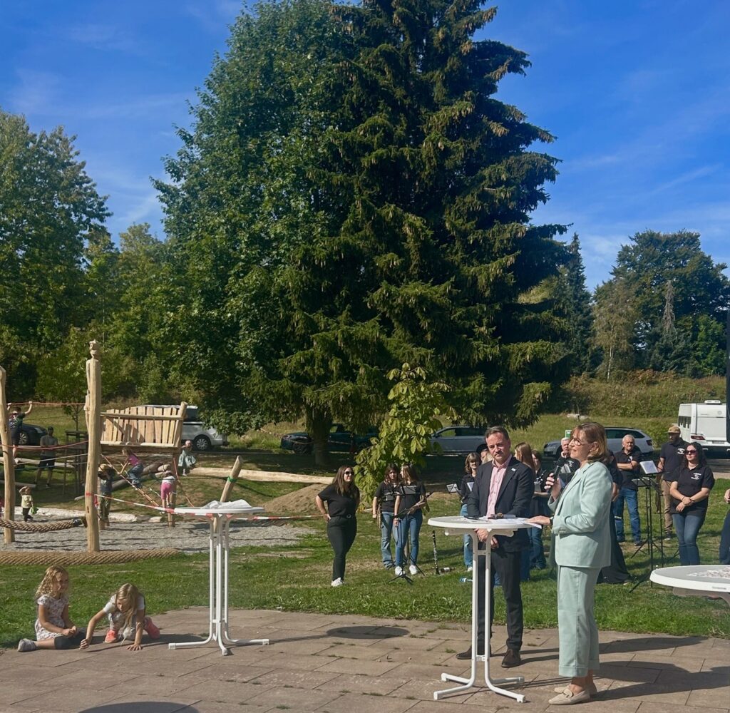Auf dem neuen Schulhof hält eine Frau hält eine Rede am Mikrophon und ein Mann steht neben ihr. Hinter ihnen befinden sich weitere Erwachsene und spielende Kinder.