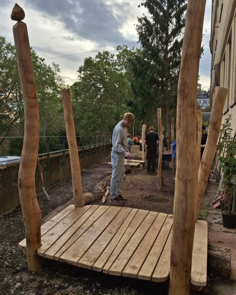 Sitzgelegenheit aus Holz, im Hintergrund steht ein Junge