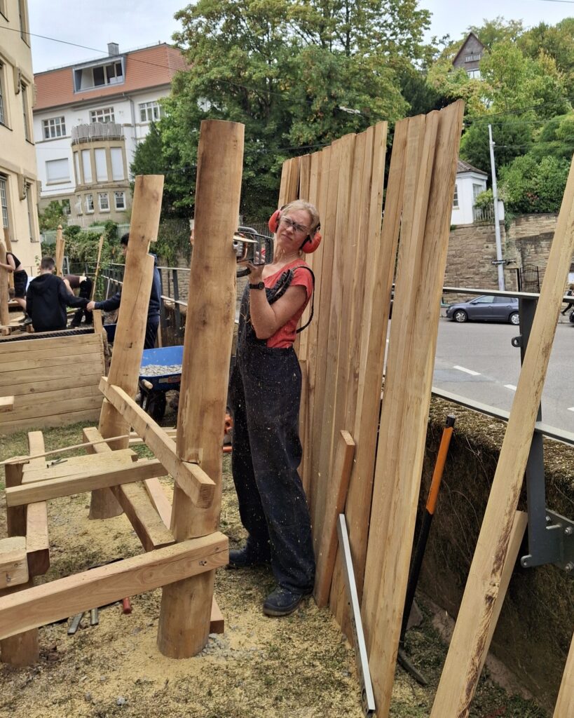 Frau bearbeitet mit Schutzbrille und Mickymäusen mit einer Maschine einen Holzpfahl.